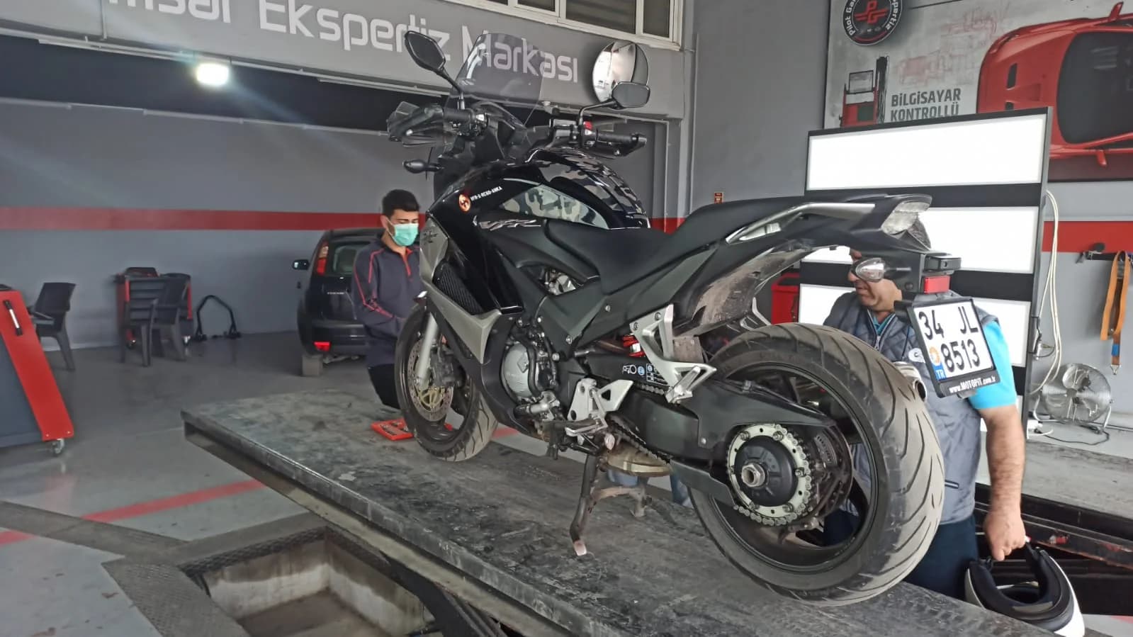 Bir ekspertiz garajında lift üzerinde incelenen siyah bir naked motosiklet ve iki teknisyen